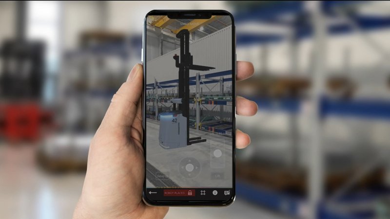 Eine Hand, die ein Smartphone vor einem Industrieregal hält. Auf dem Bildschirm sieht man ein 3D-Modell des Transportroboters auf dem realen Hintergrund